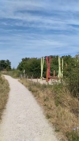 The video shows a dirt path winding through a grassy, vegetated area under a blue sky with scattered clouds. On the right side of the path, further in the distance, there appears to be some sort of playground or artistic installation with wooden elements and colorful fabric or poles. Trees and dense green foliage line the sides of the path, with dry grass and smaller plants growing closer to the path. The camera appears to be moving forward along the path, providing a first-person perspective. No people are visible in the video.