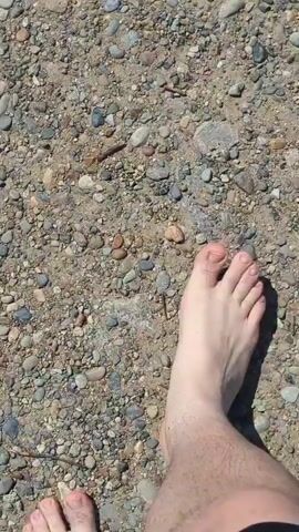 A close-up shot shows a person's bare feet stepping on a surface covered in small rocks and sand. The camera pans slightly as the person moves. The lighting suggests it is daytime and the environment appears to be outdoors.
