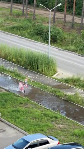 An aerial shot shows a girl on a pink bicycle riding through a large puddle on a paved path. Another person, partially visible, is on a skateboard behind her. The path is lined with green grass and surrounded by trees and a road. Several cars are parked in the foreground.