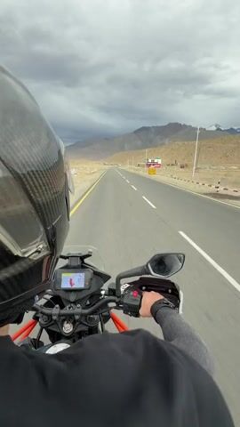 The video begins with a first-person perspective of someone riding a motorcycle on a paved road. The rider is wearing a helmet and riding gear. The road is straight and stretches into the distance, with mountains and a cloudy sky in the background. As the video progresses, the camera angle shifts, showing the motorcycle and rider from different perspectives. The rider appears to be with another person, also wearing a helmet and riding gear, who is also on a motorcycle. They are riding through a mountainous, desert-like landscape. The overall impression is of a motorcycle adventure or journey through a remote and scenic area.