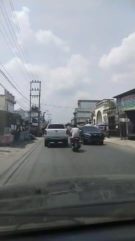 The video shows a street scene with vehicles and buildings. The camera is positioned inside a vehicle, looking forward. On the road, there are several vehicles including a white pickup truck, a black SUV, and multiple motorcycles. People are visible riding the motorcycles. The street is lined with buildings, some appearing to be shops or houses. There are utility poles with wires along the side of the road. The sky is partly cloudy, suggesting daytime. The overall impression is of a moderately busy street in a town or city.