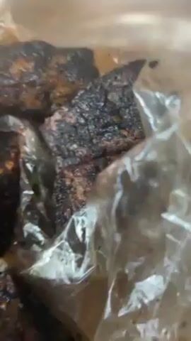 The video shows a close-up view of food in a clear plastic bag. The food appears to be cut into square pieces and heavily seasoned. The pieces are dark brown, almost black in color, possibly due to grilling or roasting. A toothpick is used to probe one of the pieces, revealing visible seasoning particles on its surface. The bag rests on what seems to be a wooden table or surface.