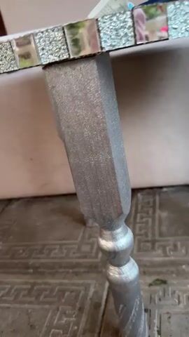 The video shows a close-up, low-angle shot of a small, round table with a mosaic-tiled surface. The table has four silver-painted legs that are turned and tapered. The legs are attached to the underside of the table with visible screws. The surface of the table appears to be made of mirrored or reflective tiles, with some colorful mosaic pieces interspersed. The table is situated outdoors on a patterned tiled floor. In the background, a light-colored, slightly textured surface, possibly a wall or a piece of furniture, is visible. The lighting suggests it is daytime, but there are no clear indicators of the time of day or weather conditions. The setting appears to be a residential area, given the tiled floor and the visible building in the background. The focus is solely on the table, with no people or other significant objects present. The video is a brief, static shot, implying it is a single capture or a very short clip. There is no visible text in the video. The overall impression is a focus on the design and construction of the table.