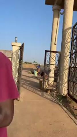 From behind a metal gate, the camera shows a person in a blue shirt, brown patterned skirt, and black head covering standing near a road. To the left, there is a large green bag with plant matter in it, and to the right, there is a broken metal object and a yellow bag. The person has their hand on their hip and appears to be looking around. The background shows a road with a white building and trees. The sky is clear and blue.