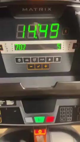 A close-up shot of a Matrix treadmill console shows a green digital display counting up time in minutes and seconds. The timer shows approximately 14 minutes and 50 seconds. Other data on the console includes a value in the 700s and the number "5." Below the display are various buttons with icons, including plus and minus symbols, and a check mark. The console has a metallic gray finish. The background is blurry, showing gym equipment, with orange elements visible. The camera is static, and there are no visible people or interactions in the shot.