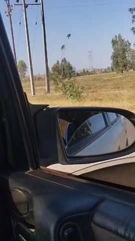 The view from inside a car shows the passenger-side rearview mirror. Reflected in the mirror is a white car, and on the side of the road, a man in a basketball jersey and straw hat is walking. The scenery outside the car window shows a rural landscape with dry grass, trees, utility poles, and a clear blue sky.