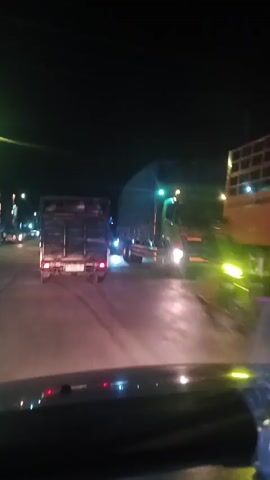 The video shows a nighttime view from inside a vehicle driving on a road. The road appears to be paved and wet, reflecting the lights of the vehicles. Several trucks are visible on the road. In the foreground, the dashboard of the vehicle the camera is in is partially visible, with some red lights illuminated. In the middle ground, two trucks are moving away from the camera. The truck on the left is smaller and appears to be a flatbed truck. The truck on the right is larger, a semi-trailer truck, covered with a tarp. The headlights of oncoming vehicles can be seen as bright streaks of light. The overall environment is dark, indicating it is nighttime. The camera appears to be held or mounted inside a moving vehicle.