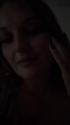 A dimly lit video shows a close-up of a woman's face. She has light skin, dark hair, and is wearing a dark top and a patterned robe. Her right hand is touching her cheek. Her eyes are looking downwards and she has a slight smile. The lighting is very low, making it difficult to see details of the environment.
