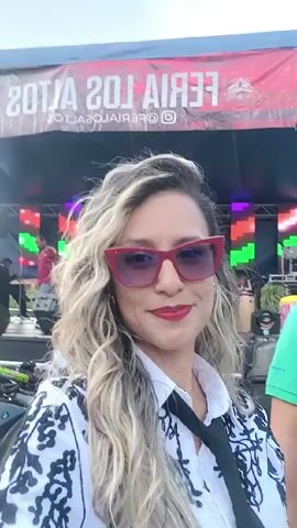 A woman in a white blouse with a black floral pattern, red sunglasses, and red lipstick, holds a camera up for a selfie. She has blonde, wavy hair. In the background, a stage is set up with equipment including drums and a large LED screen reading “MARK2”. There’s also a banner that reads “FERIA LOS”. Behind her and to the right, a man wearing a green shirt is partially visible. As the camera moves slightly, other people walk by in the background. In another shot, a large inflatable structure with the word “BERA” on it is visible, with a motorcycle parked beside it. The environment appears to be an outdoor event. There are other tents and people walking around. The day seems bright.