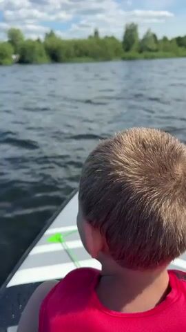 This is a close-up, over-the-shoulder shot of a young boy from behind, wearing a bright red life vest. He is sitting on a paddleboard, which is on a body of water. The paddleboard is white with grey stripes and green bungee cords. The boy's short, light brown hair is visible, with a distinct swirl pattern at the crown of his head. His right ear is also visible.

The scene is set on a lake or river, with calm, rippling water stretching out to the horizon. In the distance, a lush green tree line borders the water, suggesting a natural, possibly rural, environment. The sky above is a light blue with scattered white clouds, indicating a pleasant, sunny day, likely midday or early afternoon.

The boy appears to be looking out at the water, but his expression is not visible. The overall mood is peaceful and suggests an outdoor recreational activity, such as paddleboarding or a boat ride on a warm day. The focus is on the boy and his immediate surroundings, creating an intimate perspective of the experience. No text is visible in the media file.