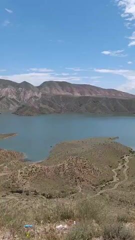 The video shows a scenic view of a large, tranquil lake surrounded by rolling, brown hills under a partially cloudy sky. The hills in the foreground are sparsely covered with vegetation and a winding dirt road leads towards the water's edge. On the right side of the frame, a concrete structure with a pipe is visible on the hillside. The lake is bordered by larger mountains in the distance.  The camera pans slightly to the right, revealing more of the landscape and the sky. A bird flies across the frame in the distance.