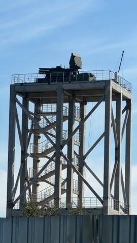 The video shows a concrete tower structure with a self-propelled anti-aircraft weapon system mounted on top. The tower has a series of stairs leading up to a platform where the weapon system is located. The weapon system appears to be a turret with a gun and a radar array. The tower is surrounded by a metal fence. The sky is clear blue.