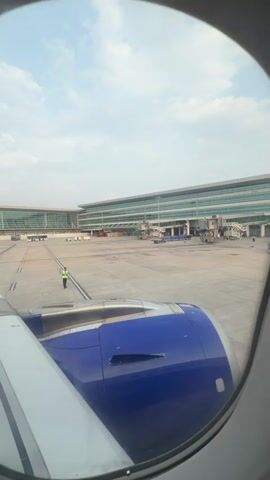 A person wearing a yellow safety vest and black pants stands on an airport tarmac, gesturing with their right hand. Behind them, a large airport terminal building with a glass facade and multiple jet bridges is visible. The wing and engine of a blue and white airplane are in the foreground, seen through an airplane window. The sky is cloudy.
