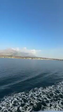The video depicts a view from the back of a moving boat on the ocean. The water is dark blue and choppy, leaving a white wake behind the boat. In the distance, there is a coastline with buildings and a mountain range, partially covered in clouds. The sky is a clear blue with a slight haze. A bird flies into the frame briefly.