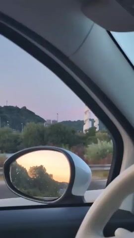 The video pans across a car interior, showing the view out the side window and the side mirror. The sky is a soft pastel color, suggesting either dawn or dusk. The side mirror reflects trees and the sky. Outside the car, two tall, white, spiral structures are visible on a hillside dotted with trees. A streetlamp is also visible. The car appears to be moving.