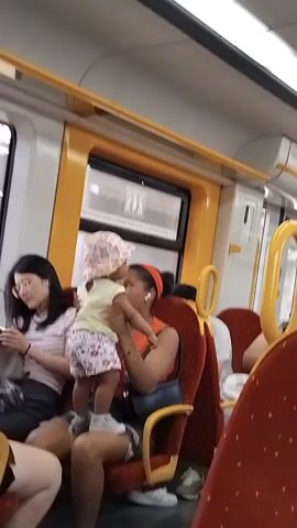 The video shows the interior of a train carriage. In the foreground, to the left, a woman in an orange tank top and denim shorts is holding a small child wearing a pink hat and patterned shorts. The child is facing the woman and appears to be interacting with her. Behind them, other passengers are visible sitting in red seats. Yellow handrails are attached to the ceiling and sides of the carriage. The overall environment is typical of public transportation.