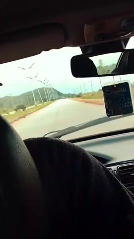 This video shows a view from inside a car driving on a paved road. The perspective is from the passenger seat, looking forward through the windshield. A dark-colored cloth is draped over the dashboard and part of the steering wheel. In the center of the dashboard, a car air freshener with the brand name "Swiss" is hanging from the rearview mirror. The road stretches ahead, with a line of streetlights on the left side, bordered by dry, reddish-brown terrain with sparse vegetation. In the distance, a few vehicles are visible on the road, and hills can be seen under a cloudy sky. The overall impression is of a journey through a somewhat arid landscape. The time of day appears to be daytime, and the weather seems overcast.