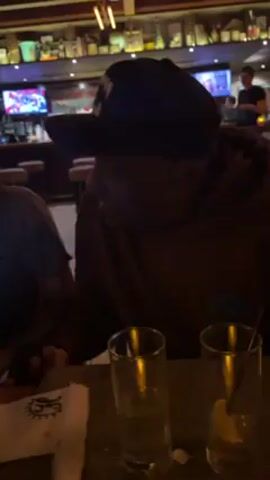 The video shows two people sitting at a table in a restaurant or bar setting. The person closest to the camera is wearing a dark hoodie and a black baseball cap. They are seated next to another individual wearing a light-colored t-shirt. There are two empty glasses on the table in front of them, one with a straw and a lemon wedge. The background features a bar area with stools, shelves stocked with bottles, and television screens displaying various content. The lighting is dim, creating a relaxed ambiance.