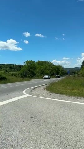 A vertical shot of a curved road on a sunny day. Several cars are driving on the road. The road is asphalt with white lane markings. There are trees and green hills in the background. The sky is blue with some white clouds.
