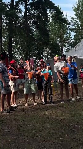 The video shows an outdoor scene with a group of people, mostly children and adults, engaged in a game. There are tall trees in the background, suggesting a wooded or park-like environment. The lighting indicates it is daytime. Several individuals, mostly children, are wearing life vests and holding orange buckets. One child in the center is attempting to catch something in their bucket, possibly a ball, as their head is tilted back and their mouth is open. Another child to their right is also looking upwards with their mouth open, holding a bucket. Two individuals, likely adults, are standing behind the children, observing and possibly participating in the game. The atmosphere appears to be casual and recreational.