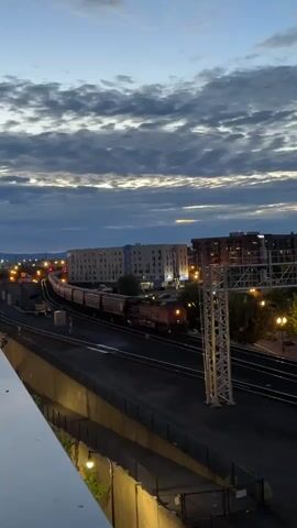 The video shows a train traveling along railroad tracks at dusk. The sky is a deep blue with dramatic clouds, and city lights are visible in the background. The train itself is long, consisting of a locomotive and multiple cars. The locomotive's headlights are on, illuminating the tracks ahead. Buildings, including a large multi-story structure, are visible on the right side of the frame. The scene appears to be captured from an elevated vantage point, possibly a bridge or overpass, as suggested by the railing in the foreground. The overall atmosphere is serene and the movement of the train is smooth and steady.