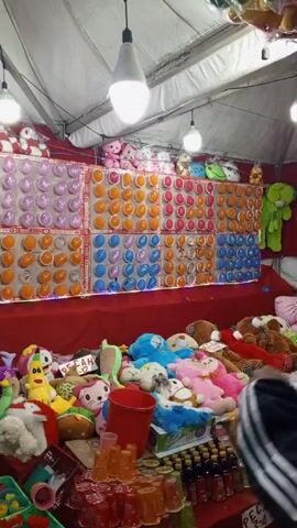 The video shows a booth at what appears to be a fair or carnival, with rows of balloons attached to a wall as a target for a game.  The balloons are arranged in a grid pattern and colored in orange, pink, and blue. Stuffed animals are placed on a table in front of the balloon wall as prizes. There are stuffed bears, cartoon characters, and other plush toys. To the bottom right, a variety of drinks and small items are visible. The booth is covered by a white tent-like structure, and a few light bulbs are hanging from above. People are present; one is seen partially behind the table on the right. The general mood is festive and the location appears to be outdoors.