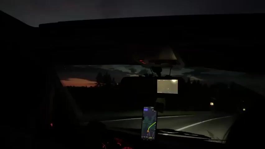 The video, shot from inside a car at night, shows the dashboard and the road ahead. A person's silhouette is visible on the left, gripping the steering wheel. In the center of the windshield, a navigation device displays a map with a green route. Above it, a small screen, likely a dashcam, shows a red-tinted image. Headlights from an oncoming vehicle are visible in the distance, illuminating the road. The sky is dark, with hints of clouds. The overall atmosphere is that of a night drive.