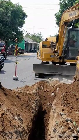 A yellow excavator is digging a trench on a dirt road. Dirt is being piled up beside the trench. The excavator has a person in the driver's seat. Behind the excavator, there are several cars, including a white van, a black van, and a silver van. There are also trees and buildings visible in the background. A red and white traffic cone is placed next to the trench. The sky is bright and appears to be daytime.