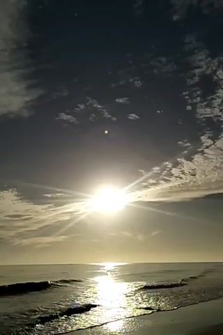 The video shows a sunny beach scene. The sun is shining brightly in the sky, with rays spreading across the scene. Some scattered clouds can be seen. The ocean is calm, with small waves gently lapping onto the sandy beach. The sun's reflection creates a shimmering path on the water's surface.