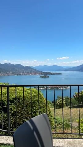 The video displays a serene, panoramic view of a large body of water, likely a lake, with mountains forming a backdrop. The sky is clear and blue with a few scattered clouds. In the foreground, a black metal railing runs horizontally across the bottom of the frame, partially obscuring a lush green hedge and a glimpse of a gray chair. Beyond the railing, the expansive lake stretches out, dotted with a few small islands and boats. On the far side of the lake, a coastal town with buildings and a cluster of trees is visible, nestled against the lower slopes of the surrounding mountains. The overall impression is one of a tranquil, scenic landscape on a bright, sunny day.