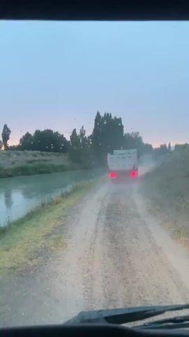 The video shows the view from inside a vehicle, looking forward and to the side. The road ahead is unpaved and dusty, with a truck driving away from the camera. To the left of the road is a body of water, possibly a canal. Trees and vegetation line the far bank of the water. The sky is overcast with a hint of dawn or dusk. A side mirror on the right shows a reflection of the road and the water, as well as the interior of the vehicle. The overall scene suggests a rural or industrial environment, possibly during early morning or late evening.