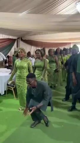 A man in a dark green, two-piece suit and black shoes dances in front of a line of people at an indoor event. The women in the line wear light green lace dresses with headwraps, and the men are in darker green outfits with vests and hats. The dancefloor has green carpet. There are tables with white linens in the background, and the ceiling is covered in white fabric. The camera follows the dancing man.