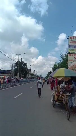 The video shows a wide paved road with a clear blue sky and white clouds overhead. Several people are walking along the road and sidewalk. On the left side of the road, there is a line of people and barriers. On the right side of the road, there are several street vendors with stalls and umbrellas. A man in a white t-shirt and dark pants is walking away from the camera down the middle of the road. Another person in a striped shirt and blue jeans is walking towards the camera on the right side of the road. A bicycle is parked next to a vendor's cart on the right. The scene appears to be a busy street in a town or city.