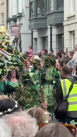 The video shows a crowd watching a parade of people dressed as the Green Man, a figure of folklore. The Green Men are covered in leaves and greenery, and some have floral crowns. The crowd is taking pictures and videos of the parade. The parade is taking place on a street in a town, with buildings visible in the background. The scene is brightly lit and colorful.