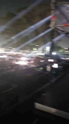 The video shows a large crowd of people at an outdoor event at night. The crowd is dense and appears to be facing a stage. Bright spotlights shine over the audience.  The background includes buildings and a tower with a red light on top. The camera seems to be positioned on a platform above the crowd.