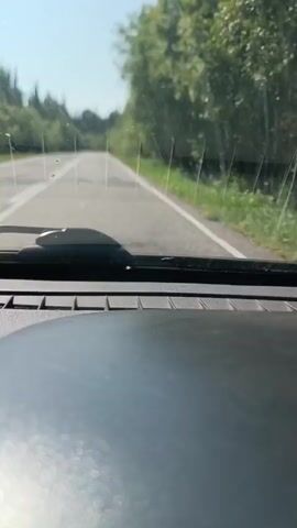 The video shows a view from inside a vehicle driving on a paved road. The dashboard is visible in the foreground. The windshield shows streaks and smudges, with a windshield wiper mechanism in the center. The road ahead is empty and appears to be in a rural or natural setting, bordered by trees on both sides. The sky is bright and clear, suggesting daylight. The camera is fixed, implying it's either mounted or held steady. The movement of the vehicle is evident from the slight motion blur of the trees and the road markings. No other vehicles or people are visible in the frame.