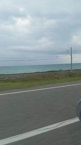 This is a side-view video taken from a moving vehicle. The view captures a shoreline with a body of water, a railway track, and a road. The water appears to be the sea or a large lake, with a slightly choppy surface under an overcast sky. A railway track runs parallel to the shore, separated by a strip of land covered in grass and gravel. Overhead power lines supported by poles are visible above the railway track. A portion of the road, with a white line marking, is in the foreground, indicating the vehicle's perspective. The overall environment suggests a coastal or lakeside route.