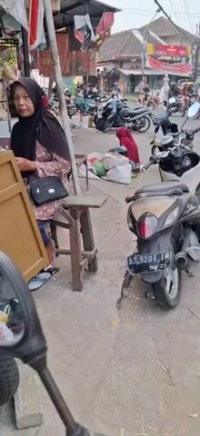The video shows a street scene, likely a market, with several motorcycles parked along the side of the road. A woman wearing a red hijab is seated on the ground next to a pile of white sacks and some green produce. She appears to be sorting or preparing the items. In the foreground, a scooter is parked, and the camera pans slightly to show more of the busy street with more motorcycles and stalls. The environment appears to be outdoors, with a paved surface and buildings in the background. There are indications of commercial activity with goods displayed and people present.
