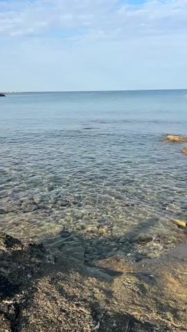 The video shows a calm sea with clear water. The seabed is visible through the water, covered in stones and pebbles. The shoreline is rocky, with some sandy areas. The sky is blue with scattered clouds. The video appears to be taken from a stationary position on the shore, looking out towards the sea. There are no people or other objects clearly visible in the scene. The lighting suggests it is daytime, likely morning or afternoon.