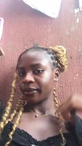 A young woman with dark skin poses for the camera indoors, against a reddish-brown textured wall. She has dark and blonde braided hair, with some braids styled in a bun on top of her head and others hanging loosely around her shoulders. She wears a black top with ruffles around the neckline and a simple gold necklace with a heart-shaped pendant. Throughout the video, she looks directly at the camera, changes her hand gestures subtly, sometimes resting her chin on her hand or touching her hair. The setting is Abakaliki, Nigeria.