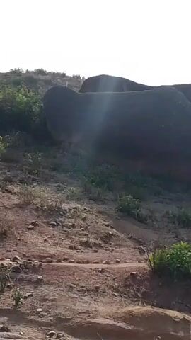 The video shows a landscape with large rocks and sparse vegetation on a hillside. The ground is mostly dirt and gravel. The sky is bright and washed out, suggesting it's a sunny day. The camera appears to be handheld, causing some shakiness. There are no people or animals visible in the frame. The landscape has a somewhat dry, arid feel to it. There is a building visible at the edge of the frame at one point.