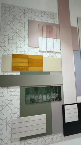 The video showcases a modern interior space, likely a design exhibition or showroom in Rho, Italy.

The initial shot focuses on a wall display composed of various tile and panel samples. The display features a white background with a subtle geometric pattern. Different sections of the wall are adorned with:
- Top left: Pink/rose gold glossy rectangular tiles and white rectangular tiles.
- Mid-left: Yellow/orange glossy rectangular tiles, below a beige/cream panel.
- Right side: Several long, vertical rectangular panels in shades of pink, beige, light grey, and light blue.
- Bottom center: A section of dark green textured glass or tile.
- Bottom left: White rectangular tiles on a grey background.
- Bottom right: White rectangular tiles on a black background.
Further along the display, visible as the camera pans, are dark blue, red, and teal square/rectangular tiles.

As the camera pans to the right, the scene widens to reveal a lounge or interaction area. Several people are present. A woman in a white shirt and black vest walks from left to right past the wall display. Another woman, dressed in a dark brown/burgundy leather jacket and dark pants, is seen bending over a long, white table, appearing to interact with an object on it. Other individuals are seated at the table, one looking at a phone, and another person is partially visible in the background. A man in a suit is also briefly visible to the far right.

The lounge area features a long, minimalist white table with water bottles and metallic bowls/dishes. The space is brightly lit by modern linear light fixtures suspended from the ceiling, some of which emit a pink glow. The overall ambiance is clean, contemporary, and professional, suggesting an event like a trade show or a design studio presentation. No discernible text is visible in the video.