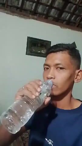 A man is drinking from a clear plastic bottle of water. He is wearing a dark blue t-shirt. The background shows a wall with a framed picture and a tiled ceiling.
