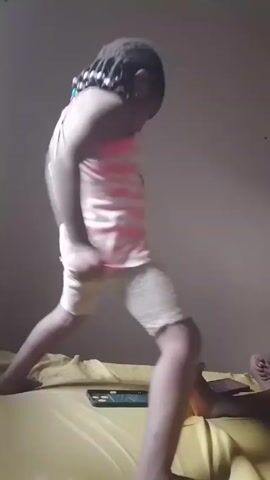 A young girl is dancing on a bed with a yellow bedsheet. She is wearing a pink and white striped tank top and light-colored shorts. Her hair is braided and has colorful beads in it. There is a mobile phone lying on the bed near her. In the bottom right corner of the video, another person is lying on the bed, with their feet visible. The background is a plain, light-colored wall. The girl is facing the camera as she dances, with movements involving her arms and body.
