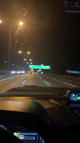 The video is shot from the perspective of a person driving a vehicle at night. The dashboard and steering wheel are visible in the foreground. The road ahead is illuminated by streetlights and the headlights of other vehicles. There are several green highway signs in the distance, indicating directions and road names. The environment appears to be a highway or a multi-lane road. The time stamp at the beginning of the video indicates 00:00.
