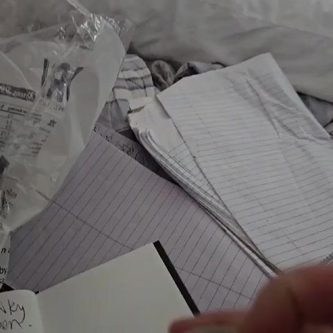 A hand is shown holding a can of V8 Spicy Hot 100% Vegetable Juice. The can has already been opened, and the hand is tapping the pull tab to make a noise. The background is a pile of miscellaneous items including paper, a plastic bag, and striped fabric.