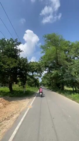 A motorbike with one rider wearing a pink shirt and a black helmet travels down a paved road bordered by lush green trees and vegetation. The sky is blue with scattered white clouds. Telephone poles with wires are visible along the side of the road. The video appears to be filmed from another motorbike following the first.