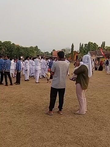 A group of people, some in white uniforms and others in casual attire, are gathered on a dry, grassy field under a pale sky. In the foreground, a man in a grey t-shirt and black pants stands facing away from the camera, while a woman wearing a hijab and a green shirt, holding a baby, stands beside him. In the background, individuals in white uniforms appear to be lining up or participating in an activity, possibly a ceremony or a school event. Several buildings and trees are visible in the distance. The overall impression is a casual outdoor gathering.