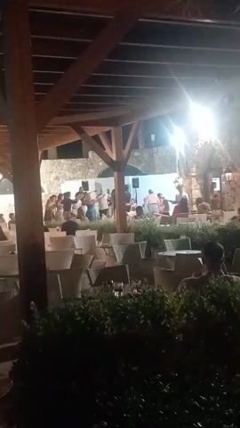 The video shows an outdoor dining area at night. There is a wooden pergola overhead with lighting fixtures. In the background, a group of people are dancing in front of a white wall. There are tables and chairs in the foreground, some empty and some occupied by patrons. Green bushes are in the lower portion of the frame.