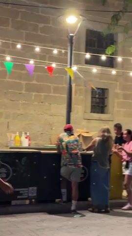 A group of people are standing around a bar area outdoors at night. The bar is decorated with string lights and colorful triangular flags. Several people are visible at the bar, some ordering drinks and others conversing. A few people are walking by in the foreground. The background consists of a stone building with windows.
